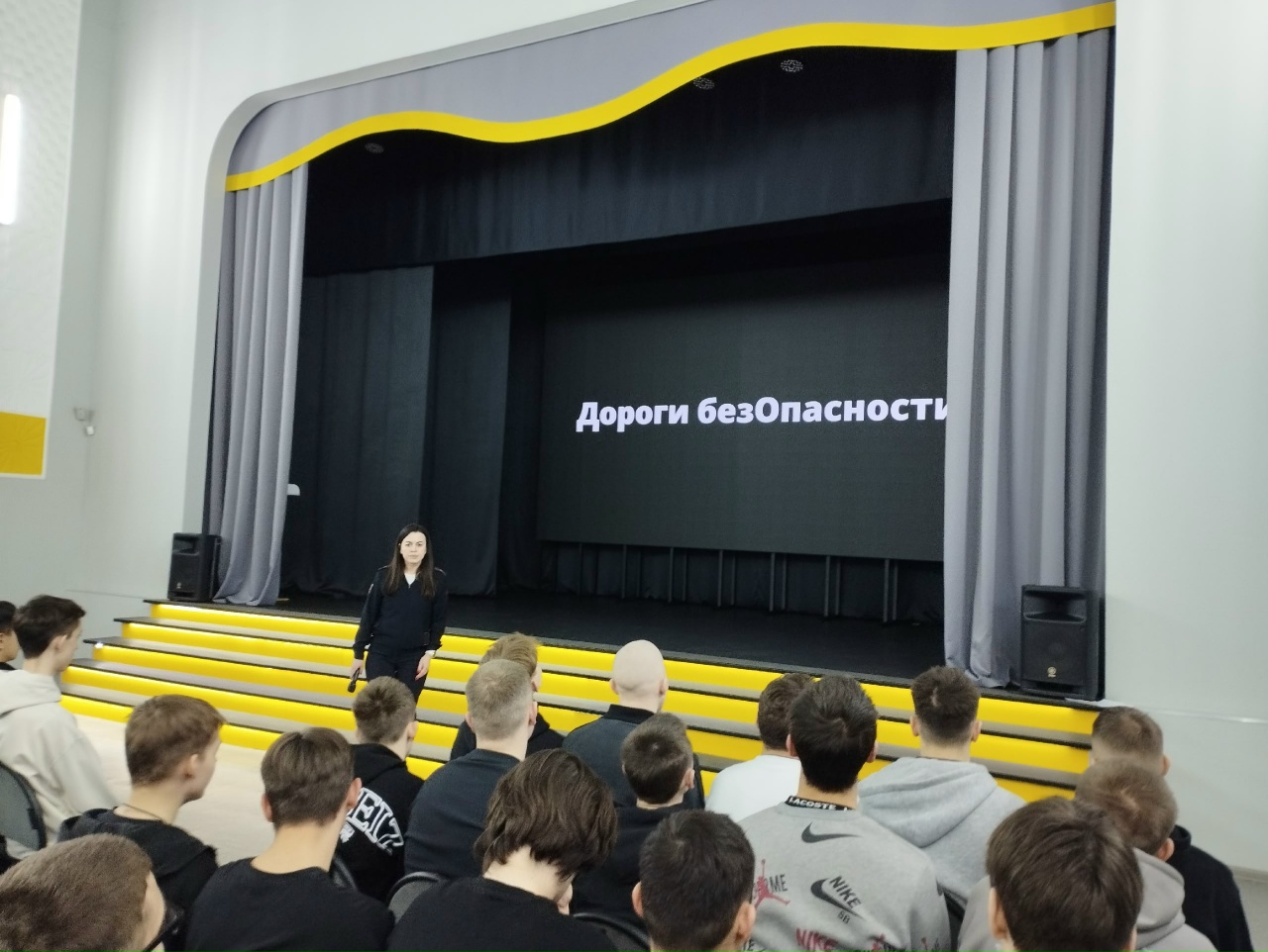 На фотографии изображена женщина, стоящая на сцене в черном костюме. Она обращается к аудитории, состоящей из молодого поколения, сидящего на желтых ступенях. На большом экране за ней написано "Дороги безОпасности". Сцена освещена, а сцена сама выглядит современно и аккуратно.