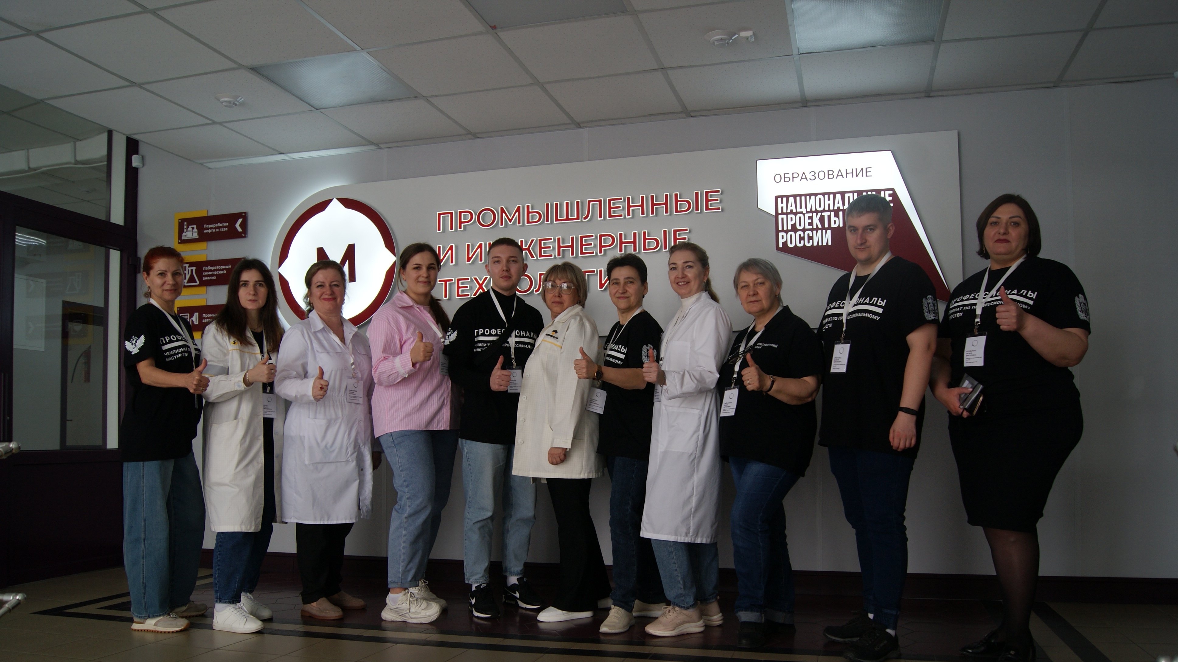 На фотографии группа из двенадцати женщин и мужчин, стоящих вместе в коридоре. Они одеты в черные футболки с надписями и белые лабораторные халаты. Все участники выглядят уверенно и выглядят дружелюбно, поочередно смотря на фотографа. На заднем плане видна светлая стена с логотипом или названием учреждения, что придает фотографии профессиональный вид.