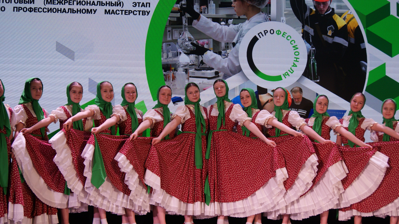 На сцене представлены женщины в традиционных нарядах. У каждой из них красное платье с белыми горошками, на головах чеканные платки. Они стоят в стройном ряду, поднимая юбки и создавая эффект волны. На заднем плане виден логотип мероприятия, а также изображения людей, занимающихся научной деятельностью. Атмосфера торжественная и радостная.