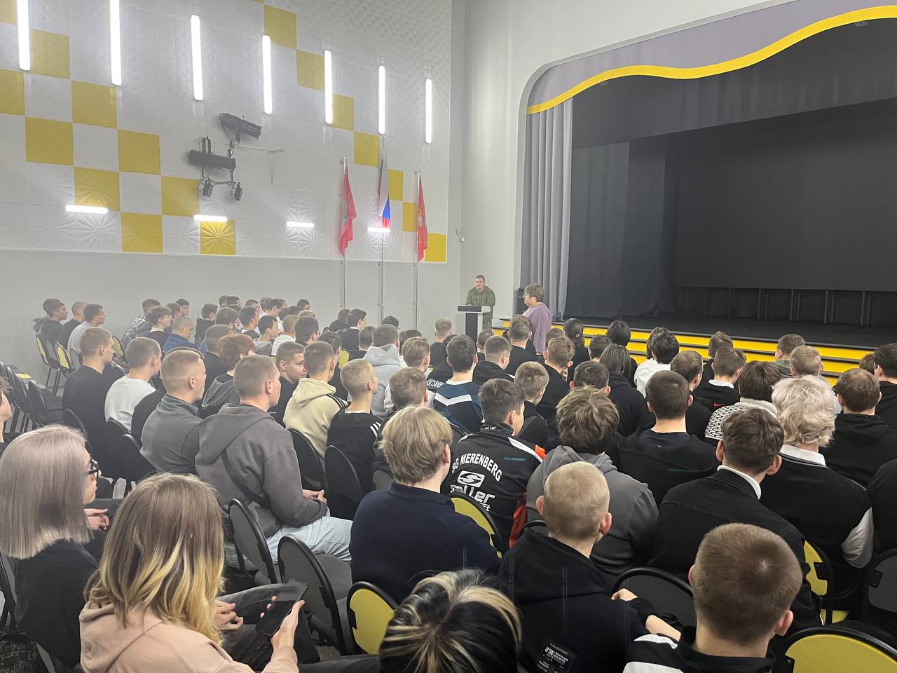 На этом фото запечатлено большое количество людей, собравшихся в конференц-зале. Зал оформлен в светлых и желтых тонах, на стенах – жёлтые квадраты. Сначала можно увидеть людей, сидящих на черных стульях, ближе к сцене стоит оратор у микрофона. Сначала зала смотрит в сторону слушателей, которые внимательно слушают. В первом ряду сидят молодые люди с различной прической, часть из которых светлыми волосами.