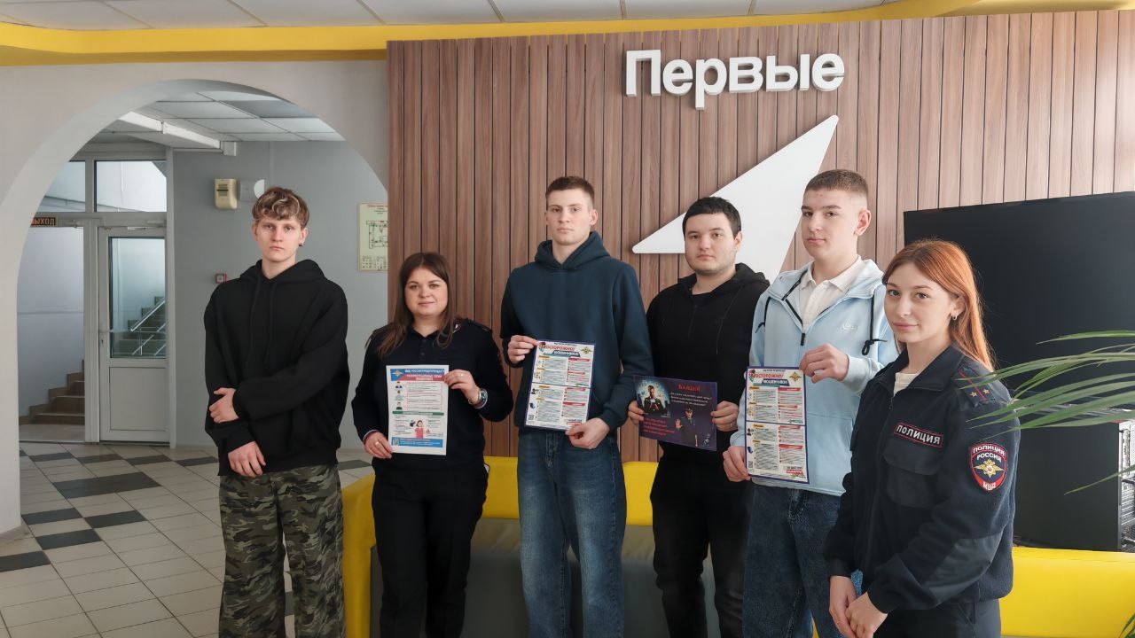 группа из шести молодых людей стоит у стены, на которой написано "Первые". Они расположены в полукруге, лицо каждого видно. У троих из них в руках листовки с информацией. Атмосфера дружелюбная и позитивная, одеты они в повседневную одежду: темные свитера и джинсы.