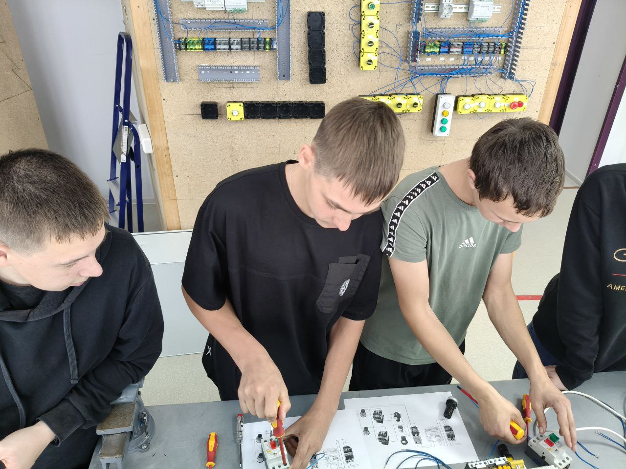 показывая троих подростков за столом. Они собирают электротехнические схемы по предоставленному чертежу, используя отвертки и различные компоненты. На стене позади них закреплены станции с кнопками и реле. Студенты сконцентрированы на процессе монтажа, заняты разбором и соединением проводов и деталей.