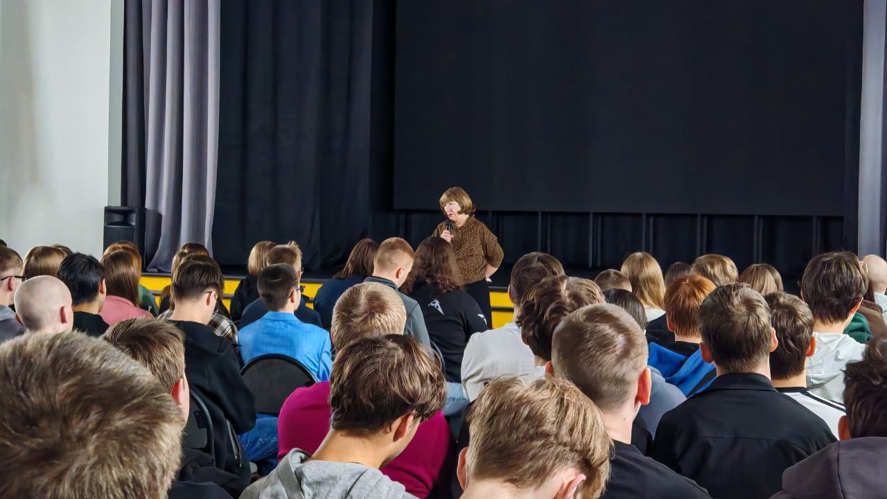 На этом снимке показано, как женщина говорит перед аудиторией, в то время как зрители находятся на своих местах, внимательно смотря на нее. Зал оформлен в светлых тонах, а на заднем плане находится черный экран. Опять же, большинство лиц в аудитории не видно, так как они находятся в профиль или со спины. Женщина одета в коричневый свитер и держит микрофон в руках, а зрители, в основном в молодежной одежде, сосредоточены на ее речи.