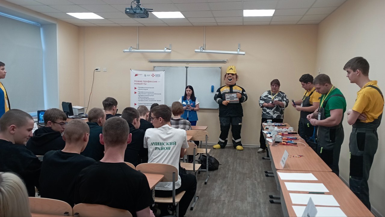 показан класс, в котором обучаются студенты. Один из них, одетый в костюм с символикой, стоит рядом с преподавателем и объясняет что-то аудитории, состоящей из молодежи, сидящей за партами. К комнате прикреплен проектор, и на стене висят обучающие материалы. Здесь фокус на взаимодействии между обучающим и учащимися, создавая атмосферу учебы и сотрудничества.