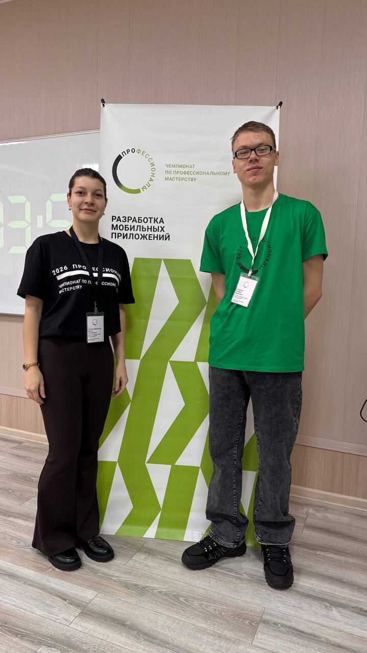 На изображении два человека стоят рядом на фоне светлого стенда. Слева — женщина в чёрной футболке с надписью на белом фоне и тёмных брюках. Справа — мужчина в ярко-зеленой футболке и очках, он выглядит дружелюбно. У обоих на шее идентификационные карточки. Внизу виден светлый пол и часть стенда с логотипами.