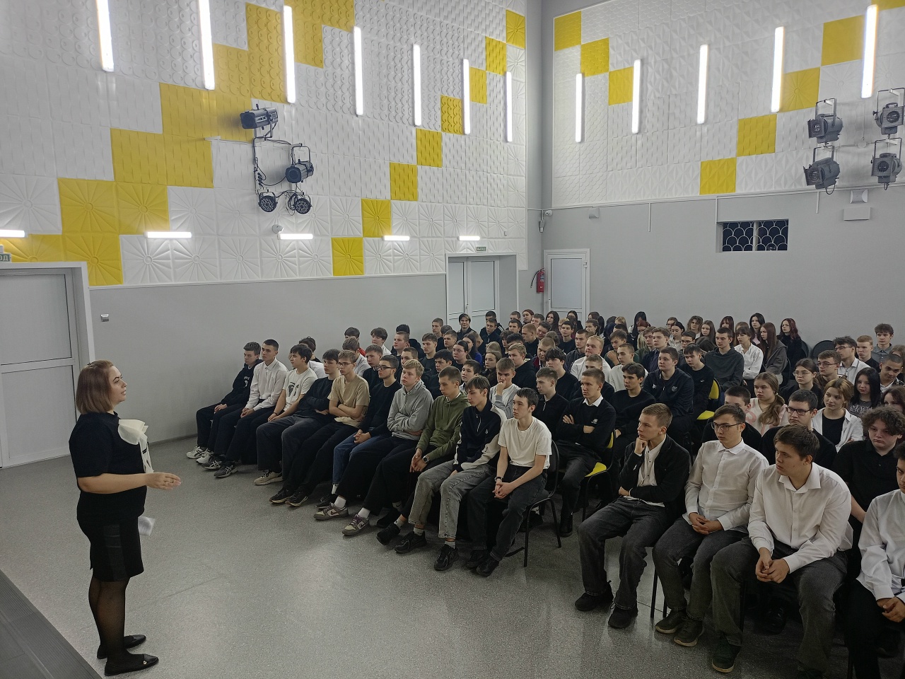 запечатлён момент, когда спикер стоит перед группой студентов и проводит часть семинара или лекции. Обстановка может быть более непринужденной, а студенты, располагающиеся в аудитории, выглядят заинтересованными и вовлечёнными в обсуждение. Форма одежды студентов варьируется, что создает неформальную атмосферу. Лектор, вероятно, делится практическими советами или личным опытом, что подчеркивает значение такого рода мероприятий для студентов.