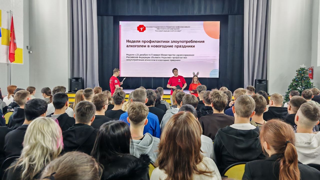 изображена сцена мероприятия, где выступают три человека. На экране за ними демонстрируется презентация с текстом на тему борьбы с зависимостями. В зале сидит множество слушателей, большинство из которых занимаются спортом. Помещение оформлено флагами, а в правом углу виден украшенный новогодний ель. Аудитория внимательно слушает выступление.