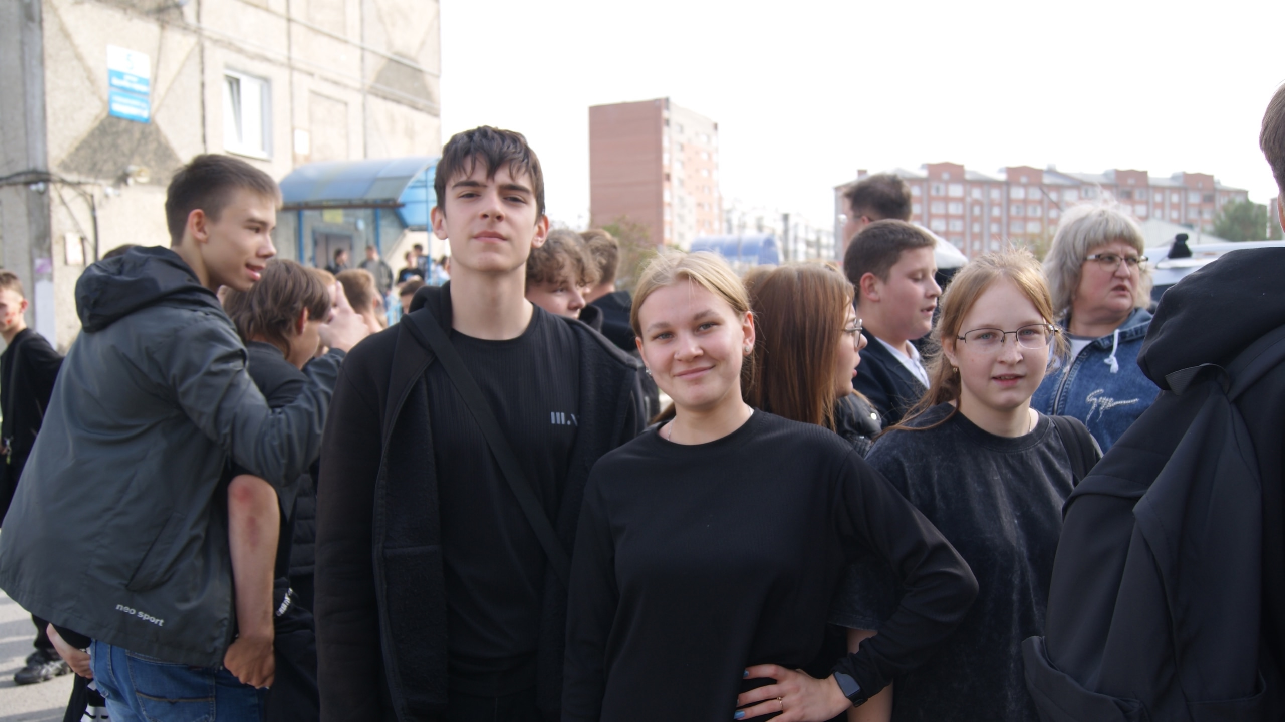 На фотографии три подростка стоят на фоне группы людей. Два из них — парень и девушка — улыбаются и смотрят в камеру, оба одеты в тёмную одежду. На заднем плане видны другие молодые люди, некоторые из которых также одеты в чёрное. Сзади можно разглядеть здания и автобус.