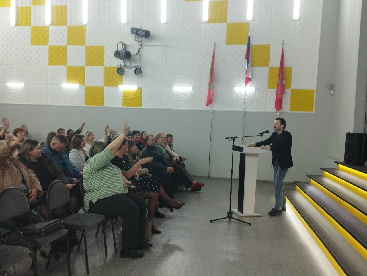 момент активного участия зрителей. Человек на сцене говорит в микрофон, а часть аудитории подняла руки, явно взаимодействуя с ним. Зал оформлен в том же стиле, что и на предыдущих фото, с желтыми квадратами на стенах. Аудитория включает людей различных возрастов, а сцена выделяется простым черным пьедесталом.