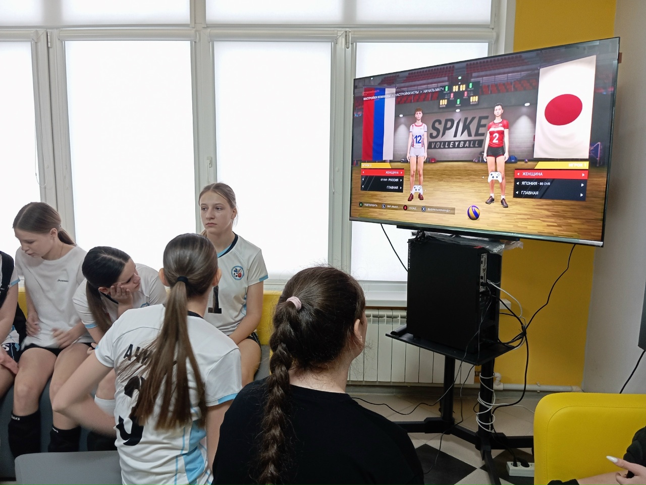 фотография показывает сцена, на которой несколько девушек сидят рядом с экраном, на котором отображается игра в волейбол. На экране видны данные о командах – одна из них представляет Россию, а другая – Японию. Обе команды изображены в форме, соответствующей их странам. Девушки, находящиеся на переднем плане, смотрят на экран, некоторые из них обсуждают происходящее, а другая девушка с длинными волосами сидит и внимательно наблюдает за матчем. Интерьер, как и на предыдущей фотографии, яркий и современный.