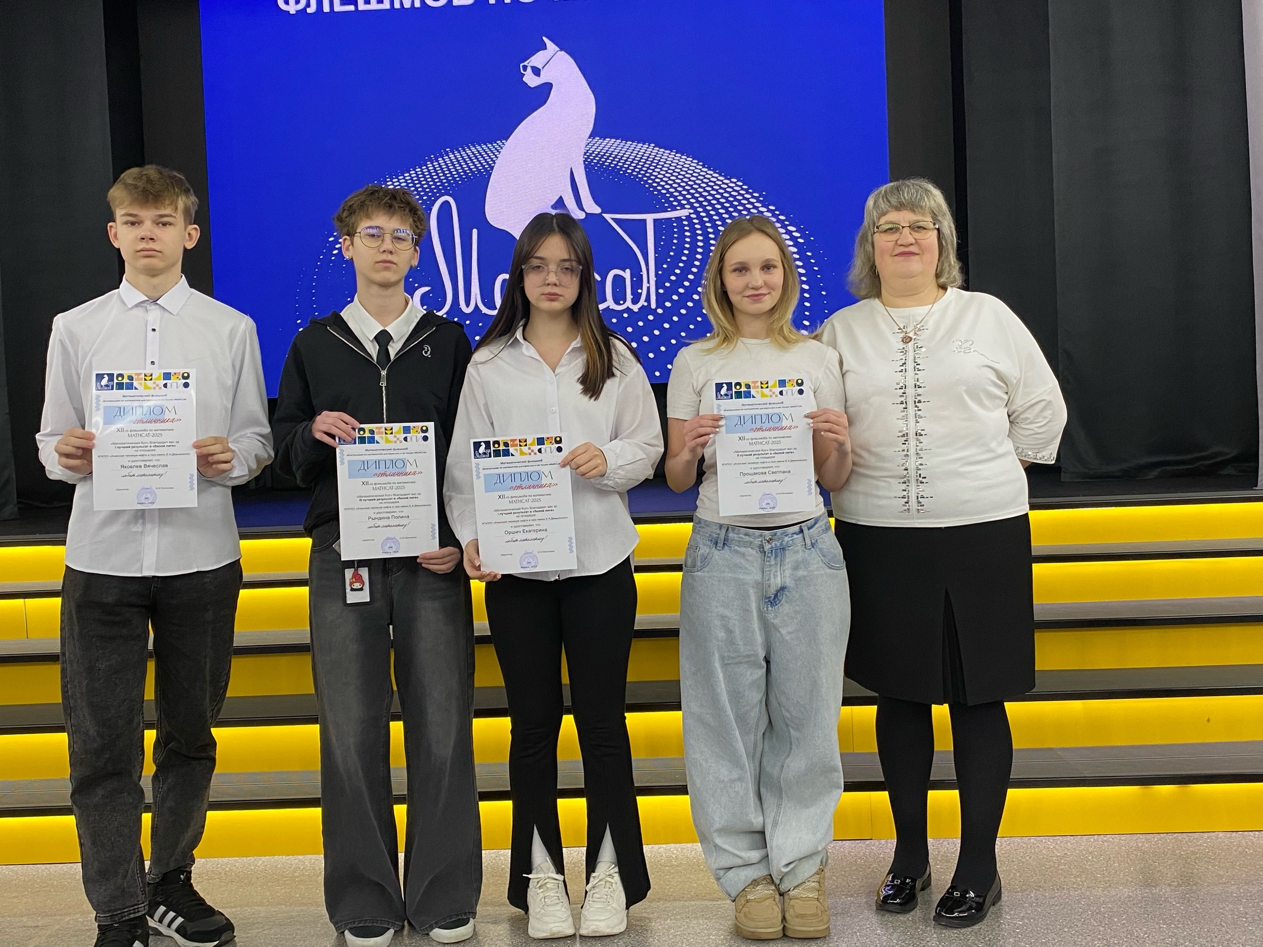 На сцене пятеро человек. Слева стоит молодой человек в белой рубашке и черных брюках, держащий диплом. Рядом с ним - девушка в белой рубашке с длинными рукавами и джинсах. Далее - ещё одна девушка с длинными светлыми волосами, она также в белой рубашке и держит диплом. С правой стороны - женщина в светлой блузке, которая стоит на фоне с синем логотипом мероприятия и держит диплом. Позади них расположено светящееся оформление.
