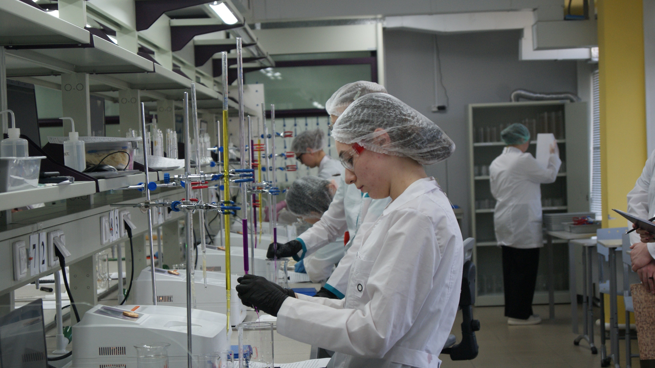 Лаборатория: несколько людей в белых халатах и сетчатых шапочках работают за длинной лабораторной скамьёй с пробирками, трубками и приборами; на переднем плане женщина в перчатках аккуратно наливает жидкость в лабораторную посуду.