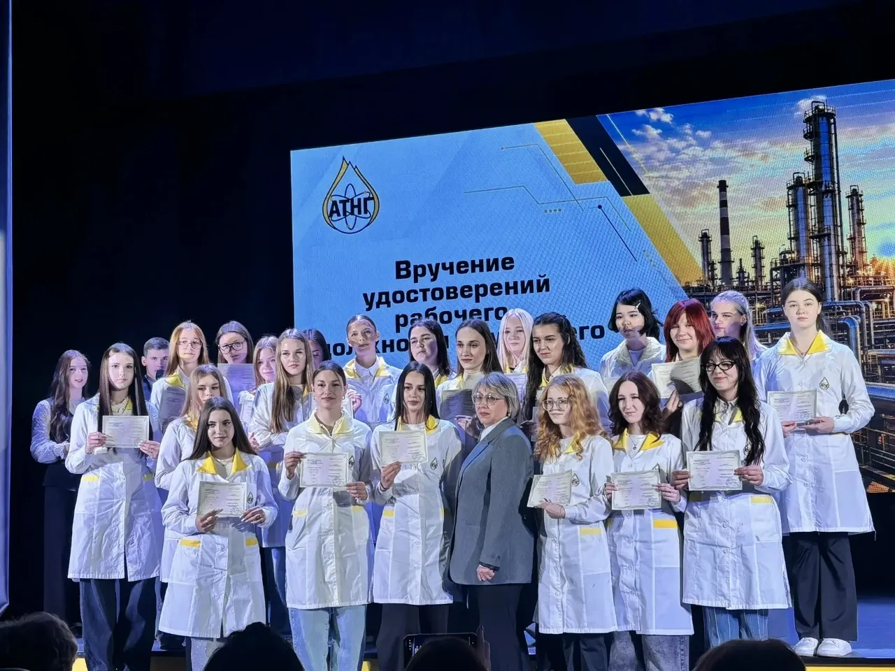 Групповое фото на сцене. Большая группа студентов (преимущественно девушки) в белых лабораторных халатах с желтыми вставками держат в руках полученные сертификаты. В центре группы стоит женщина в сером деловом костюме. На заднем плане расположен большой синий экран с логотипом «АТНГ», частичной надписью «Вручение удостоверений рабочего...» и изображением нефтеперерабатывающего завода на закате.