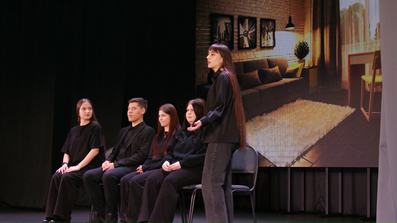 пять человек в чёрной одежде сидят на стульях на сцене. Один человек стоит, а остальные внимательно слушают. На экране за ними изображение уютной комнаты с диваном и дизайнерскими элементами. Атмосфера выступления сосредоточенная, участники выглядят увлечёнными процессом.