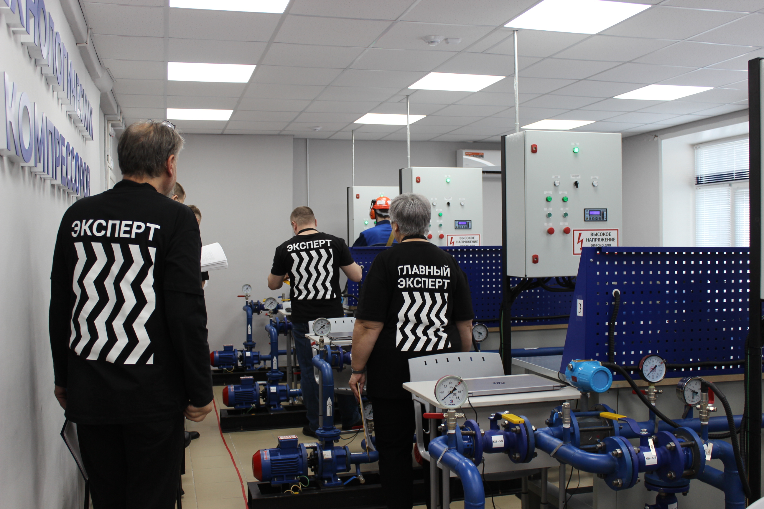 На этом снимке изображена обучающая комната с оборудованием для работы с насосами. Группа людей, одетых в черные футболки с надписями "ЭКСПERT" и "ГЛАВНЫЙ ЭКСПERT", движется к технологическому оборудованию. В комнате установлены трубопроводы и пульты управления, на которых отображаются манометры и переключатели. Атмосфера работы здесь более динамичная и целеустремленная, чем в лаборатории.