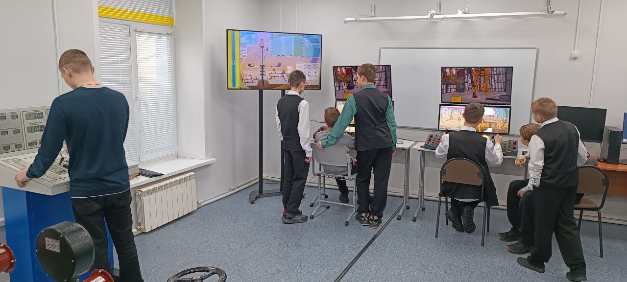 изображены подростки, стоящие и сидящие за столами с компьютерами. У них есть доступ к нескольким экранам, на которых одновременно отображаются разные симуляции. Один из мальчиков использует сенсорное устройство. Комната хорошо освещена, с большими окнами, через которые попадает свет, создавая комфортную атмосферу для обучения.