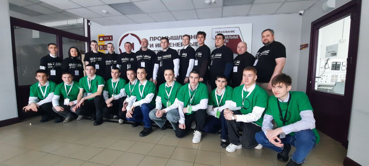 представлена большая группа из 18 человек, стоящих в помещении с нейтральными стенами. Многие из участников одеты в зеленые футболки, а некоторые — в черные. Они улыбаются и выглядят радостно. В переднем ряду сидят 6 человек, остальные стоят позади. На стене расположена вывеска, похожая на ту, что была на первом фото, но менее четкая, и текст на ней полностью не виден.