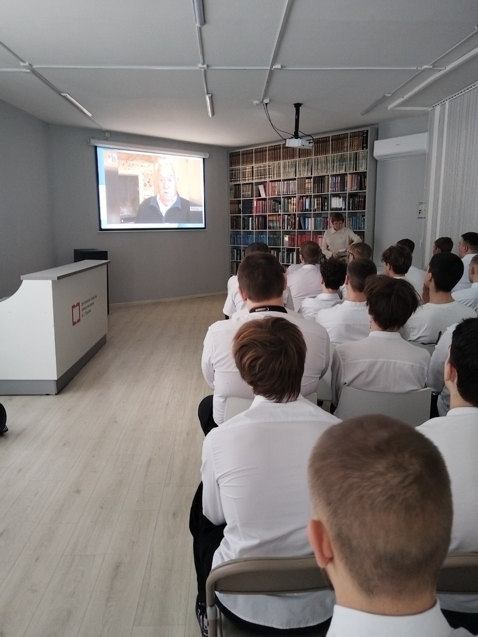 показан зал, где сидят молодые люди, вероятно, студенты. Они одеты в белые рубашки и внимательно смотрят на экран, на котором транслируется видео. На экране изображен пожилой мужчина, который, судя по всему, говорит или ведет лекцию. В зале светлые стены и пол, а справа от зрителей расположена стена с книгами.