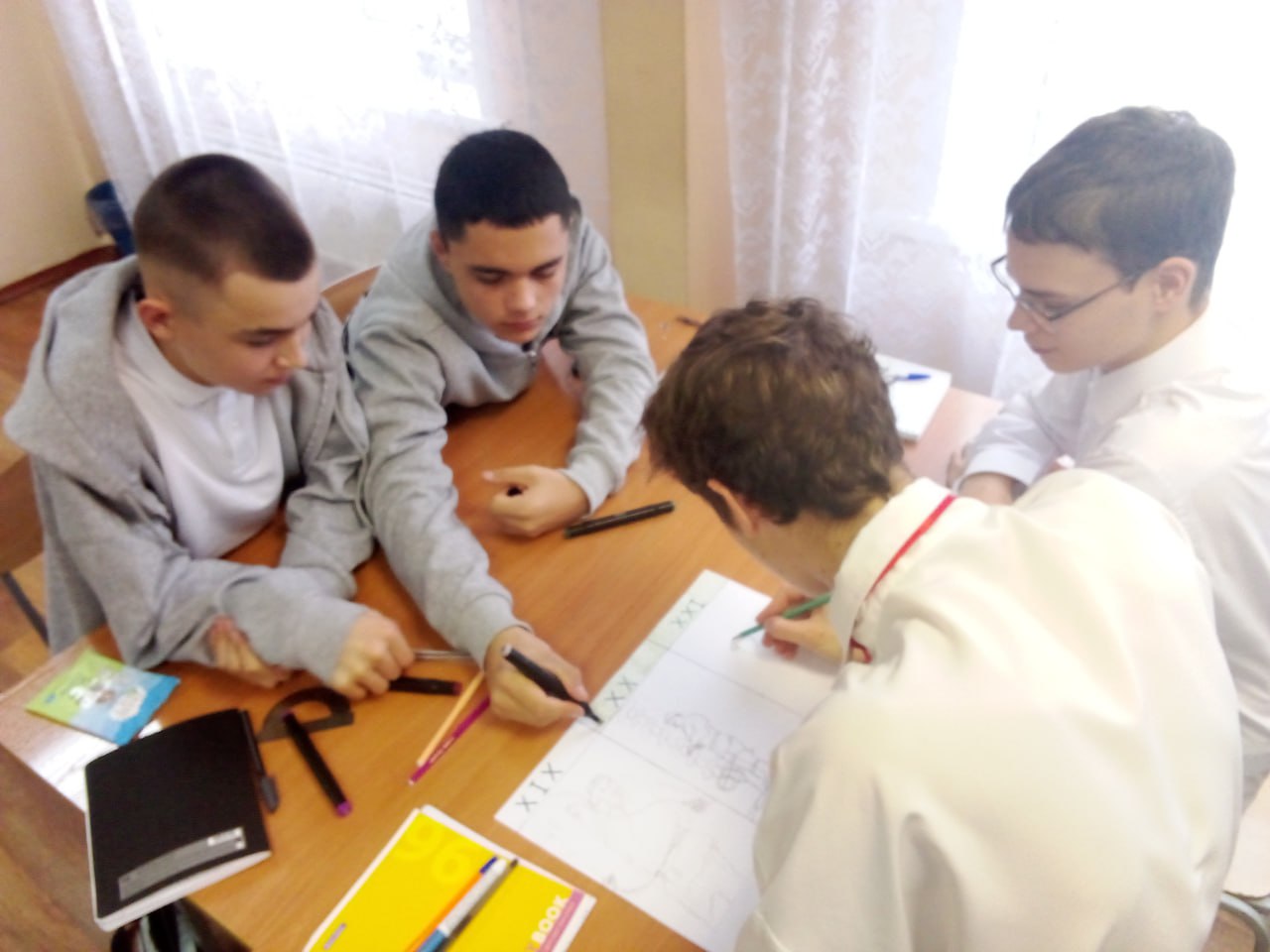 На этой фотографии изображены четыре подростка, работающих в классе: Три из них сидят за столом, сосредоточенно смотря на что-то, что находится на столе. Четвертый подросток стоит, объясняя или рисуя на большом листе бумаги, расположенном на столе. Освещение в комнате естественное, а светлые обои в сочетании с подоконниками создают дружелюбную атмосферу учебного процесса.