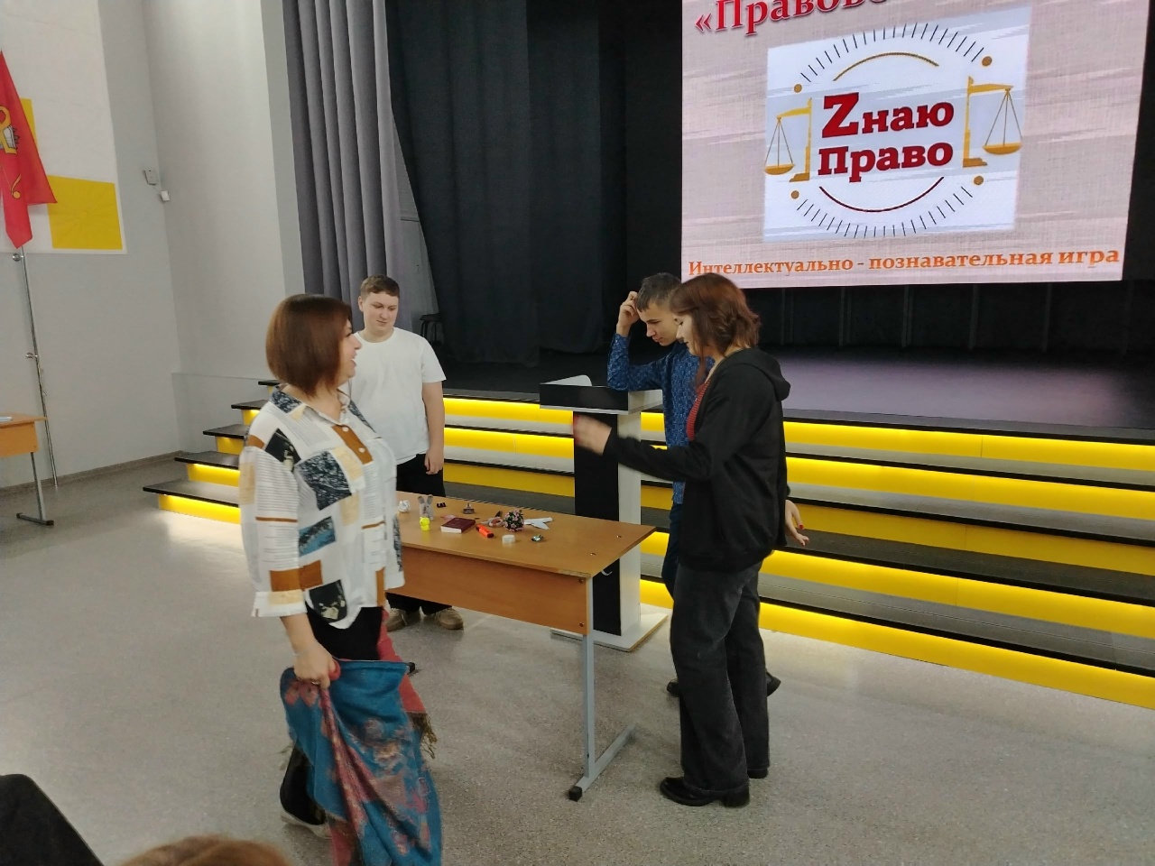 изображена сцена с людьми, стоящими возле кафедры в зале с ярким освещением. На заднем плане видно экран с надписью «Знаю право» и символикой, вероятно, связанной с правоведением. Один человек, возможно, ведёт презентацию или обсуждение, в то время как другие участники, включая женщину в свитере, слушают и наблюдают за происходящим. На столе также находятся небольшие предметы аналогичные тем, что были на первой фотографии.