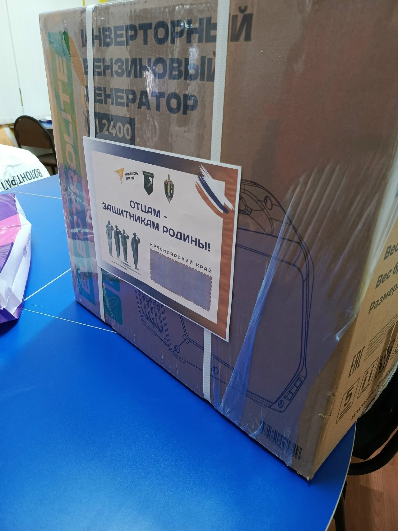 На изображении изображена коробка, обернутая в пленку и с наклейками. На коробке видно изображение инверторного генератора, а также текст: "Отцам - защитникам Родины!" Коробка размещена на синем столе. Эта фотография придает ощущение готовности к отправке или вручению подарка, подчеркивая военную или патриотическую тематику.