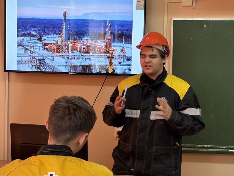 На изображении присутствует группа людей на техническом заседании или обучении, связано с энергетическим сектором. Один из участников рапорта, выполняя роль лектора, одет в защитную каску оранжевого цвета и черную рабочую куртку с желтыми вставками. Он активно жестикулирует, объясняя что-то важное. На фоне, на экране, изображение нефтяной платформы в окружении леса и гор, что указывает на тематику лекции. Другие участники, сидящие за столом, повёрнуты к лектору, что подчеркивает их внимание к информации. Классная доска на стене добавляет образовательную атмосферу.