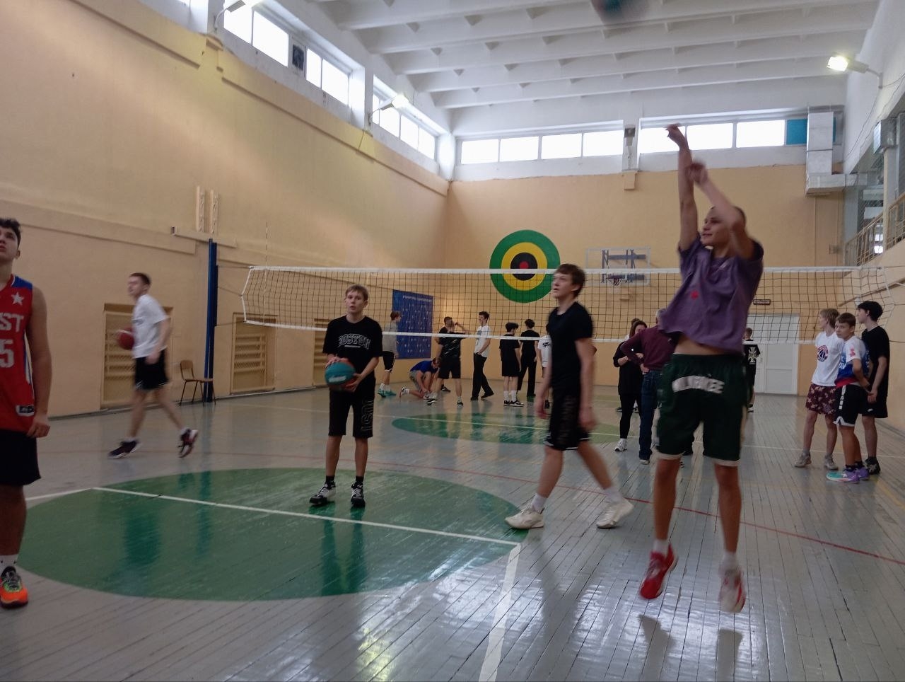 На фото изображена волейбольная площадка в спортивном зале. В центре сцены находится группа подростков, играющих в волейбол. Один из мальчиков присел и готовится к выбросу мяча, находясь в воздухе, в момент удара. Остальные ребята распределены по площадке, некоторые из них стоят вокруг сетки, а другие подбирают мяч. Пол зала зеленый с серыми полосами, а стены оформлены в светлых тонах с округлыми мишенями для метаний.