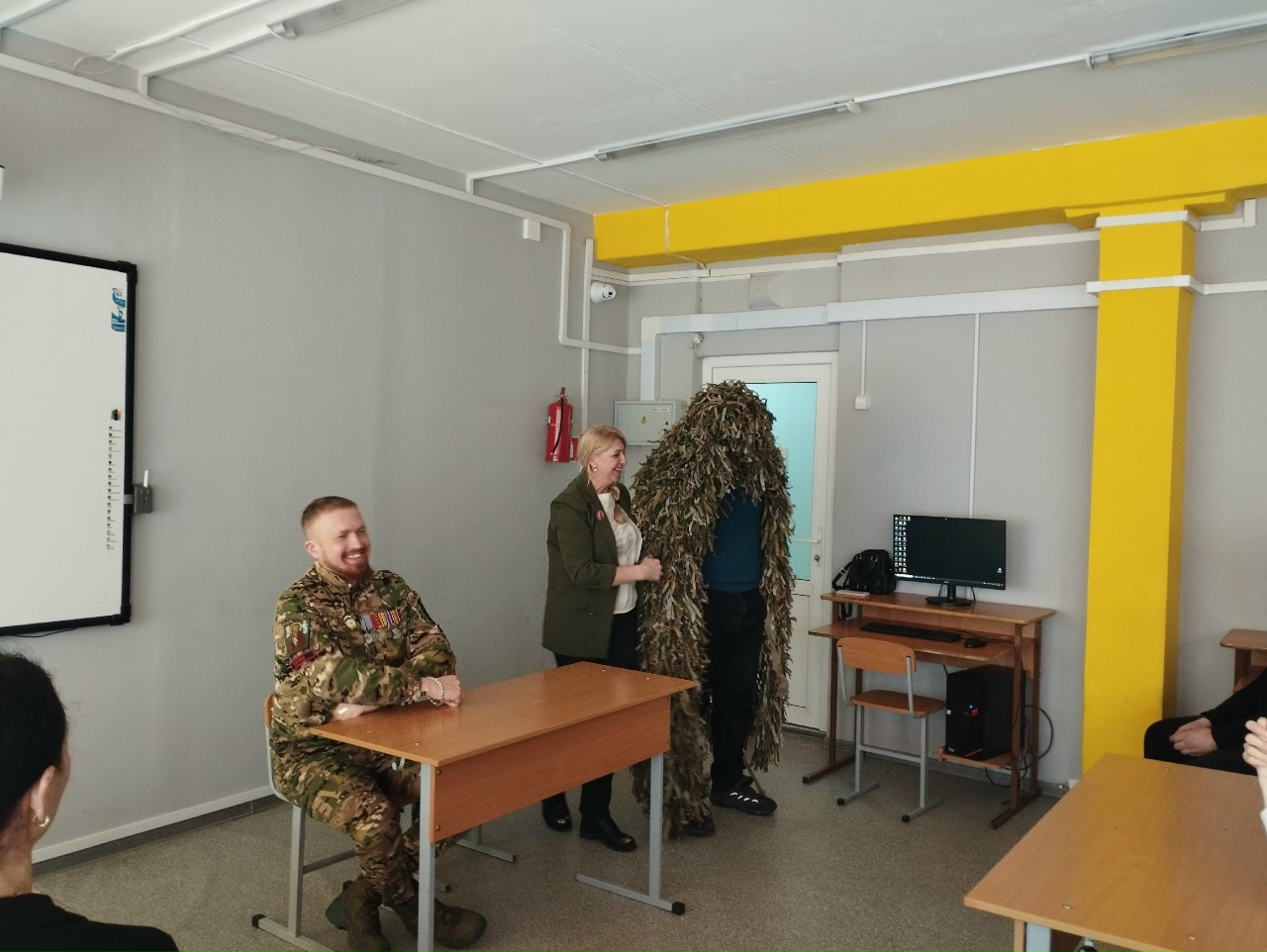 На фотографии мужчина в камуфляжной форме и женщина в деловом костюме сидят за столами в учебном кабинете. Женщина, кажется, говорит, а рядом с ней стоит человек в маскировочном костюме, который выглядит комично и необычно. В помещении есть освещение и компьютер на столе, создающее атмосферу учебной обстановки.