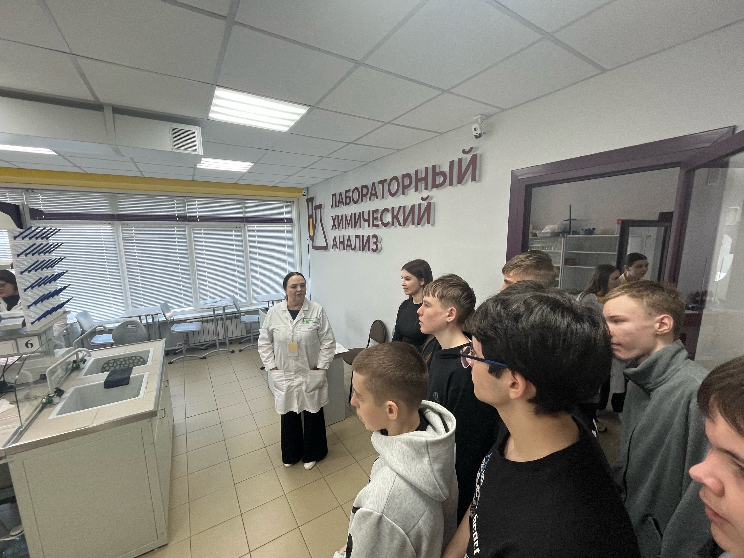 представлена группа подростков, смотрящих на женщину в белом лабораторном халате, которая объясняет что-то у лабораторного оборудования. Фон занимает исследовательская лаборатория с современным оборудованием. Стены и окна оформлены в светлых тонах, освещение яркое. На заднем плане видно название лаборатории, написанное крупными буквами. Участники группы выглядят заинтересованными, слушают attentively.