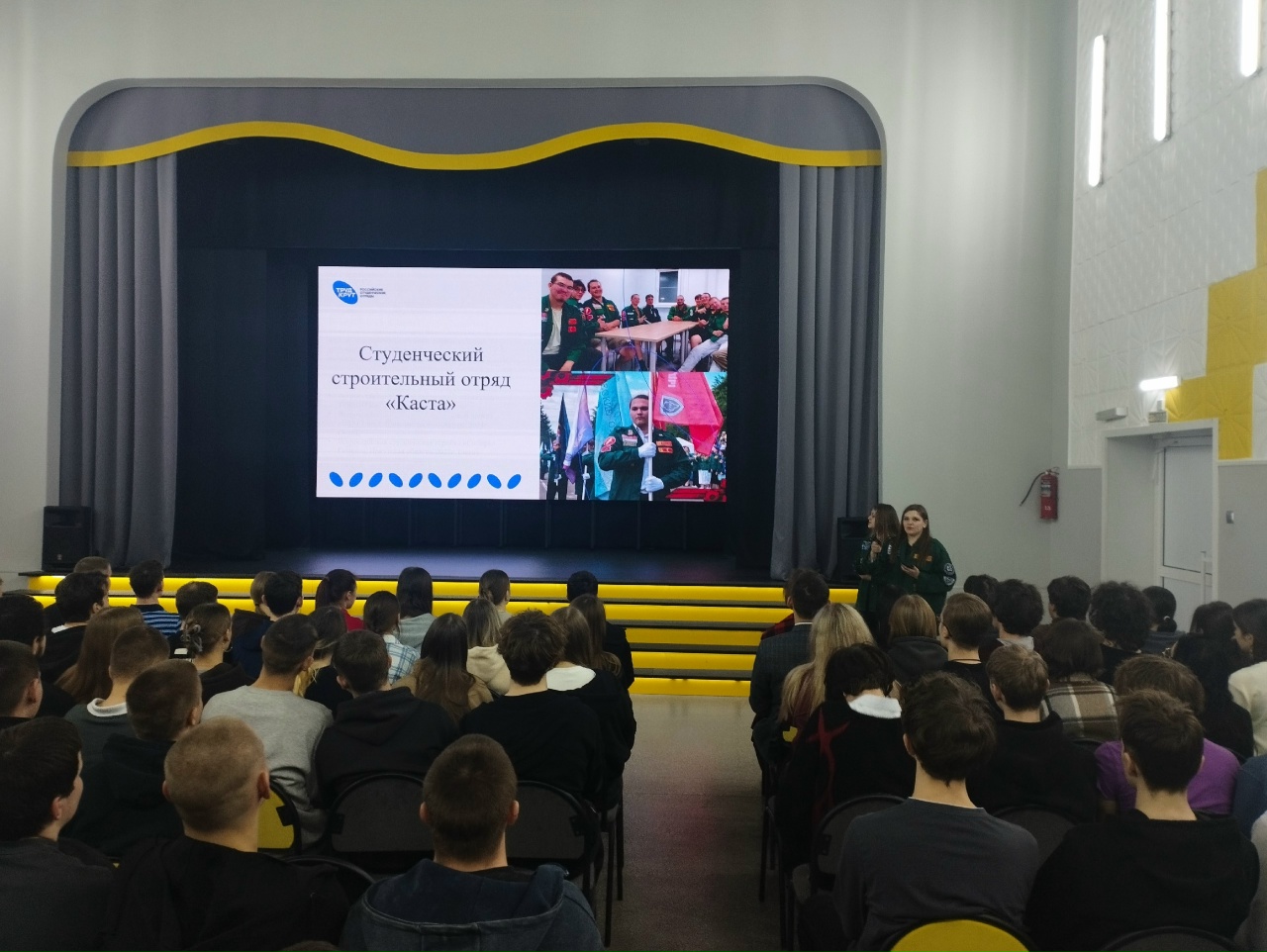 обширный зал, в котором сидят множество людей. Они повернуты к большой экранной панели, расположенной в фокусе. Экран показывает информацию о студенческом строительном отряде с названием "Кастю". На экране можно увидеть фотографии участников в форме и, возможно, различные символы или логотипы, относящиеся к отряду. Зал оформлен в светлых тонах с серыми элементами и жёлтой рамкой по нижнему краю экрана, придающей ему современный вид.