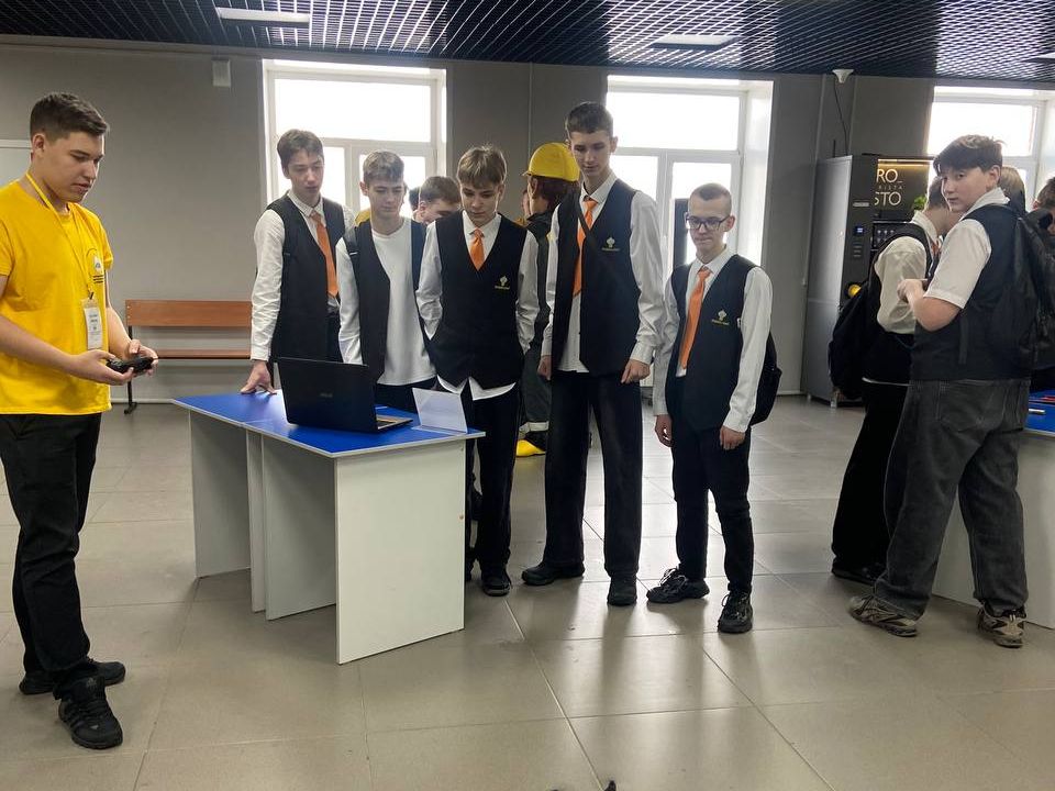 На фотографии группа юношей стоит возле стола с ноутбуком. Один из участников, одетый в яркую жёлтую футболку, объясняет что-то остальным, которые внимательно его слушают. Все они в формальных нарядах: чёрные жилеты и оранжевые галстуки. В помещении есть светлая отделка и стеклянные стены, что делает обстановку более современной. За ними видны рекламные баннеры и выставочные стенды, что указывает на проведение какого-то мероприятия или выставки.