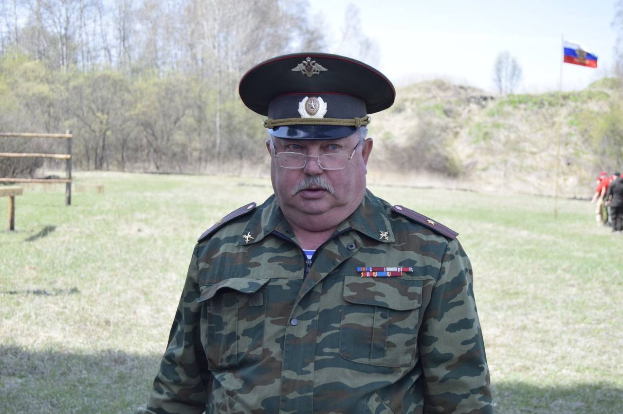 На первом фото изображён мужчина средних лет в военной форме. Он носит гимнастерку с камуфляжным узором и фуражку, украшенную гербом. Лицо серьёзное, на нём очки, а на груди располагаются награды и знаки отличия. На заднем плане виднеется зелёная трава и светлые деревья, а также флаг России, развевающийся на ветру.