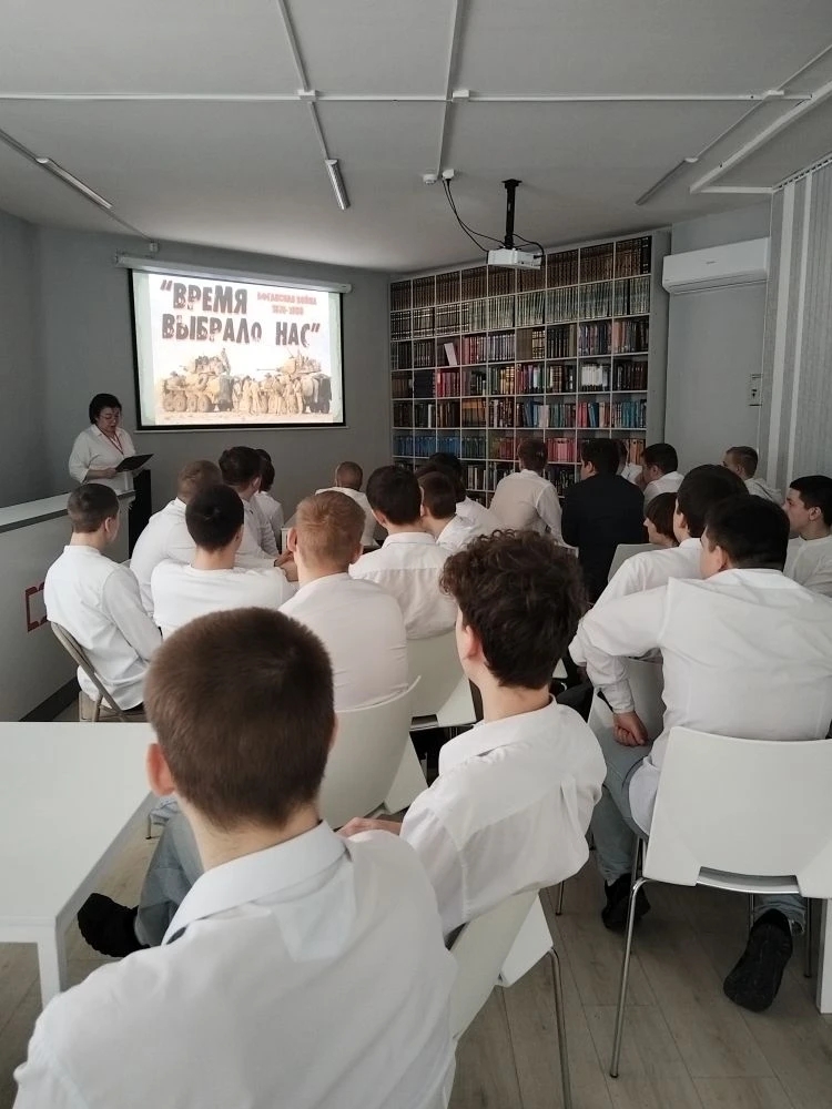 Третье изображение показывает занятие в аудитории, где также видно множество студентов в белых рубашках. Перед ними выступает женщина с визуальным материалом на экране, на котором написано «Время выбрало нас». На стенах можно заметить полки с книгами. Обстановка выглядит сосредоточенной, все участники внимательно слушают.