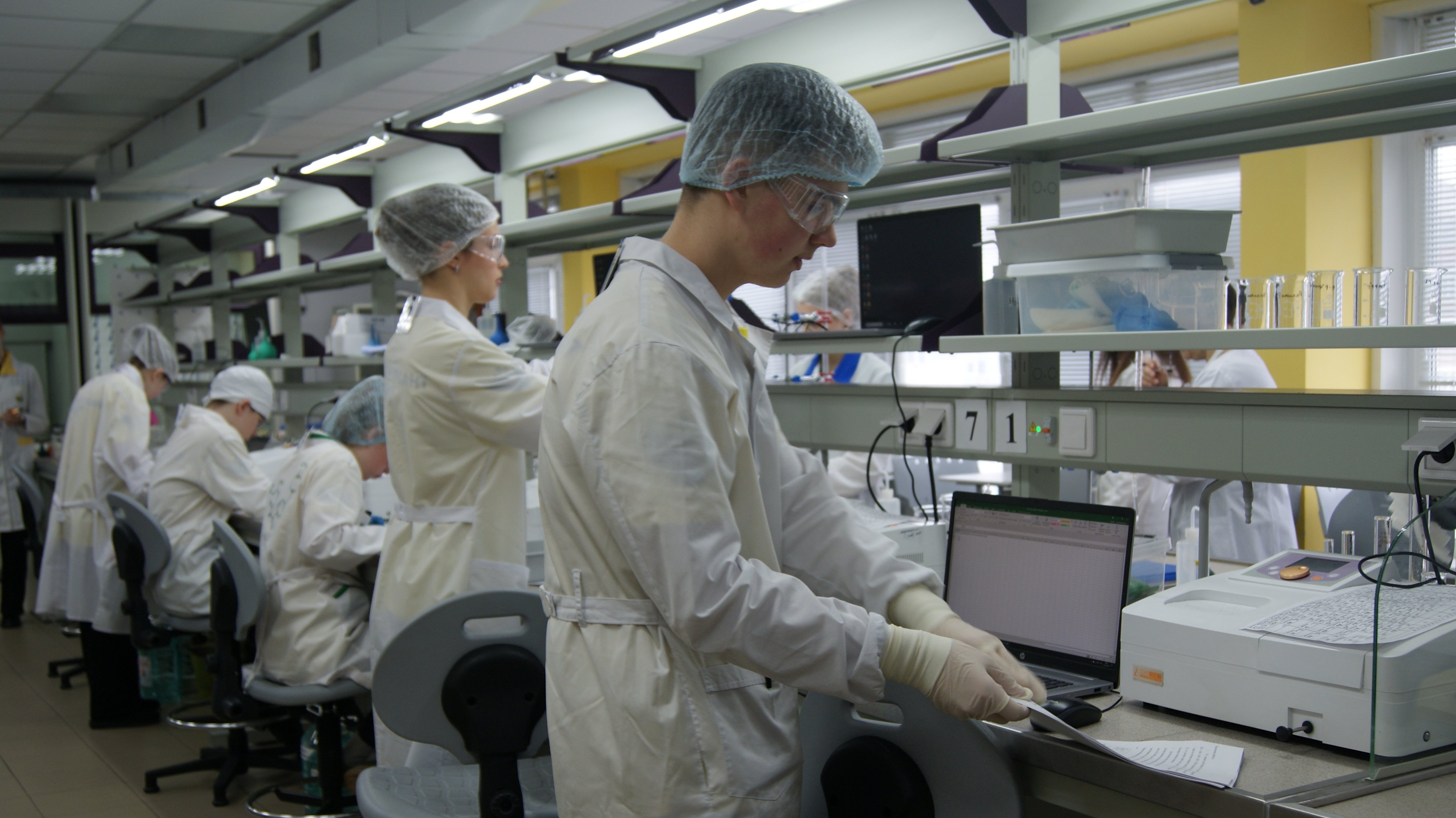 В лаборатории несколько ученых работают над научными проектами. В центре изображения молодой человек в белом лабораторном халате и защитных перчатках стоит у рабочего стола с ноутбуком. Он сосредоточен на своей задаче и держит в руках инструмент для работы. На заднем плане находятся другие сотрудники, также одетые в халаты и головные уборы, которые занимаются своими делами.