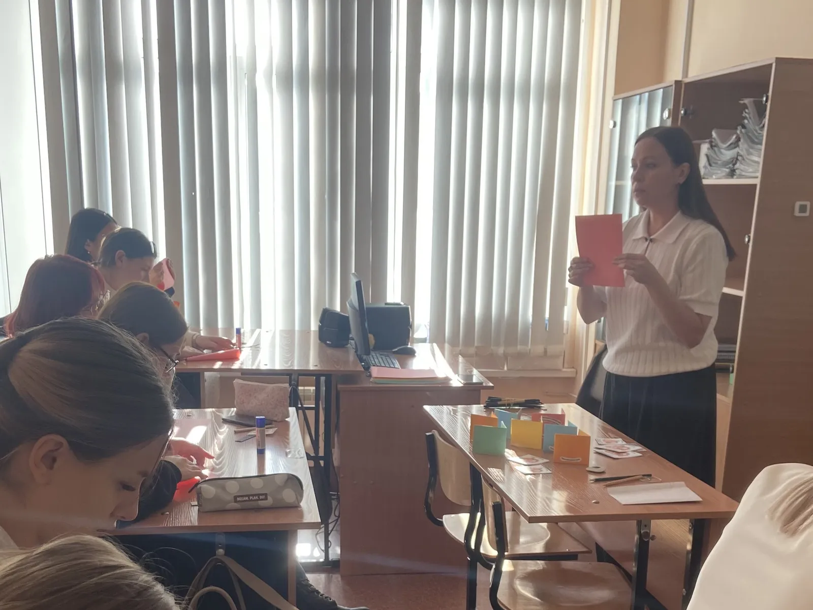 Женщина-преподаватель стоит у окна с вертикальными жалюзи и держит в руках красный лист бумаги, обращаясь к ученицам. Девушки сидят за столами, на которых разложены разноцветные карточки, ручки и пеналы. Справа стоит шкаф с бумагами.