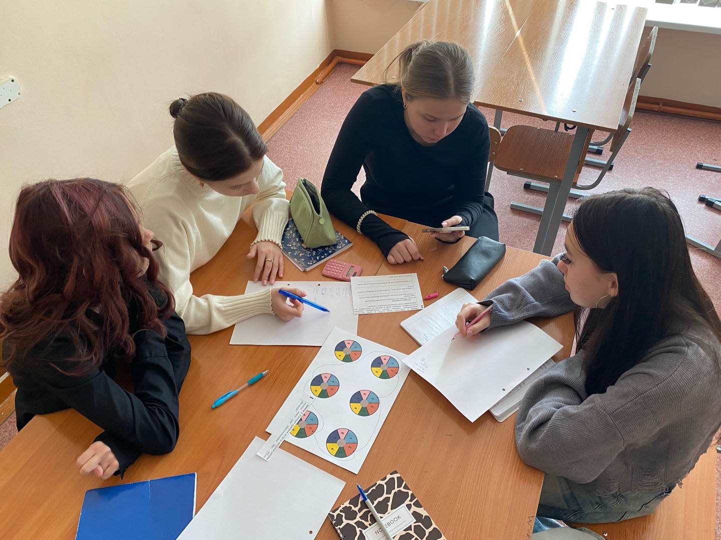 четыре девушки сидят за столом в похожем помещении, сосредоточенные на своей работе. Каждая из них имеет блокнот или лист бумаги. Одна девушку одета в белый свитер, другая в черный. Они обсуждают информацию, а на столе лежит несколько распечаток с цветными кругами и заметками. Как и на первом фото, они выглядят заинтересованными и вовлеченными в процесс.