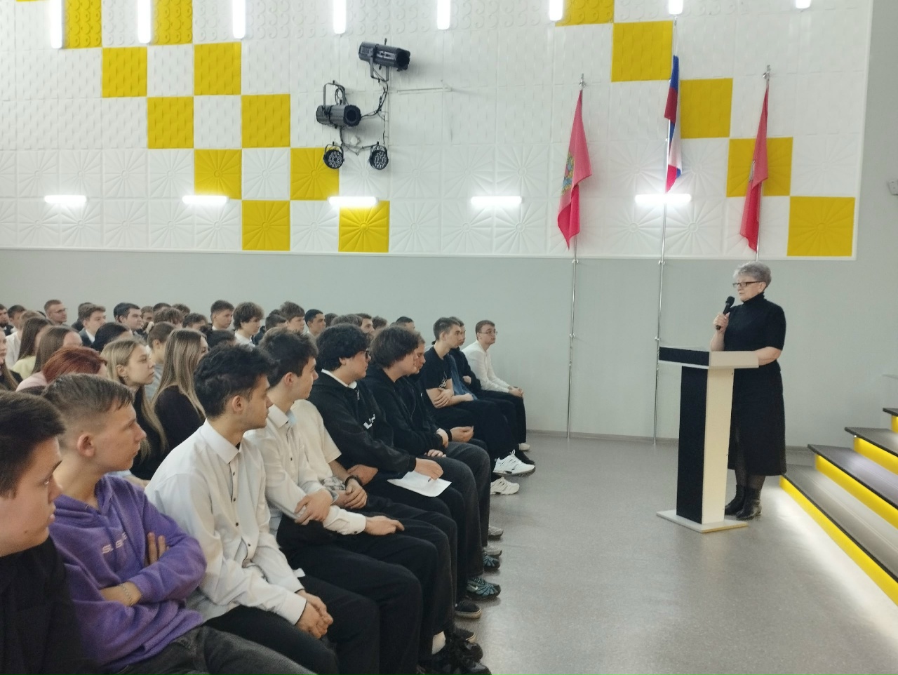 Аудитория: ряд молодых людей (студентов) сидит в первом ряду, плотно друг к другу. Большинство повернуто вправо и внимательно смотрит на сцену; у некоторых в руках блокноты или папки. Одежда повседневная — свитеры, рубашки, тёмные брюки. Слева на стене декоративный узор из жёлтых и белых квадратов; рядом установлены три флага на пилонах. Освещение обычное, обстановка формальная, аудитория спокойна и сосредоточена.