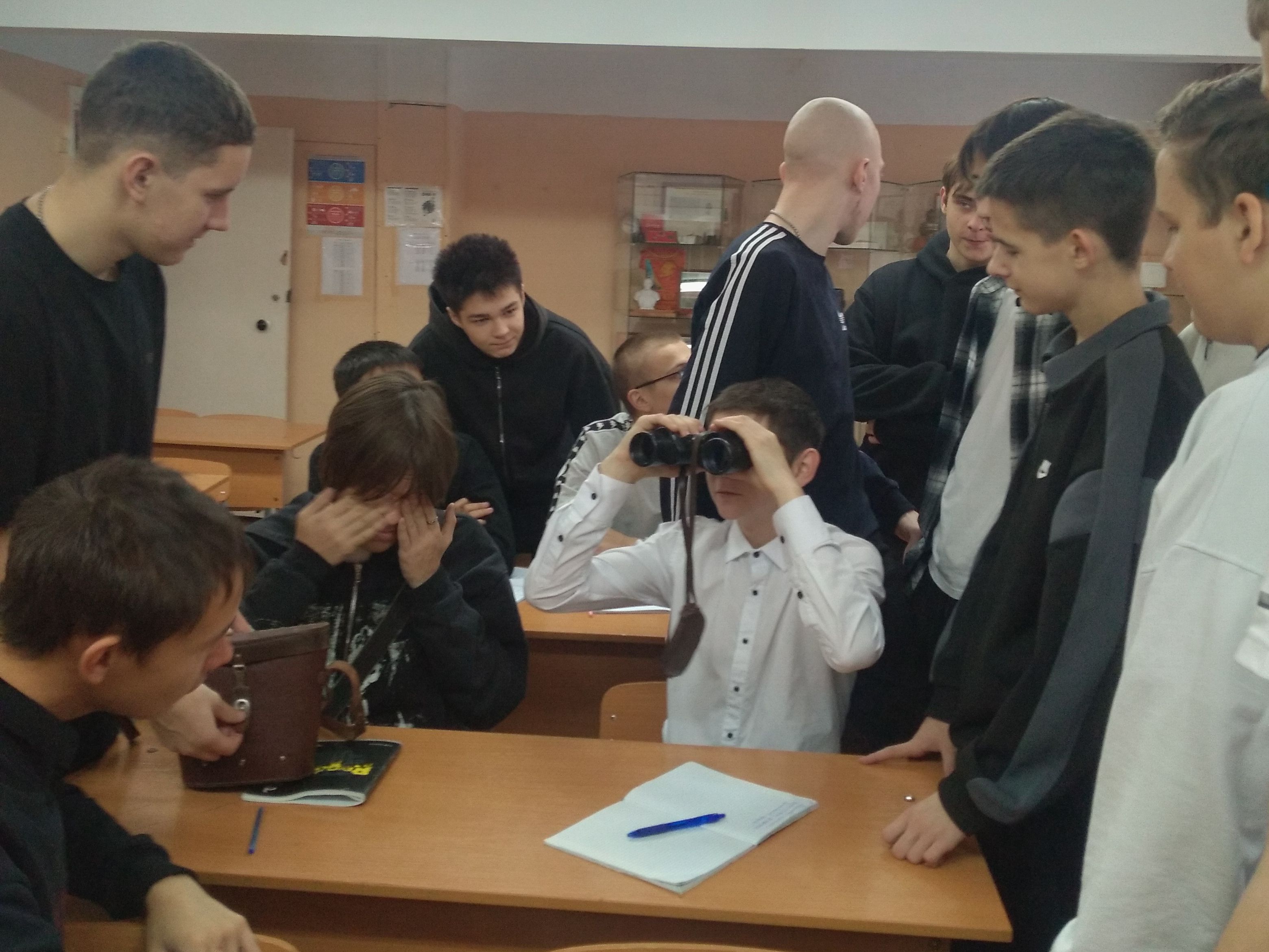 На этом фото группа подростков, собравшихся в классе. Один из мальчиков стоит с биноклем, который прицеливает в группу своих сверстников. Некоторые из учеников, стоящих рядом, смеются или с интересом наблюдают за тем, что происходит. В центре внимания — мальчик, который закрывает глаза, а также несколько других ребят, склонившихся к столу, где лежит тетрадь и ручка. Освещение в классе достаточно яркое, а атмосфера выглядит дружелюбной и веселой.