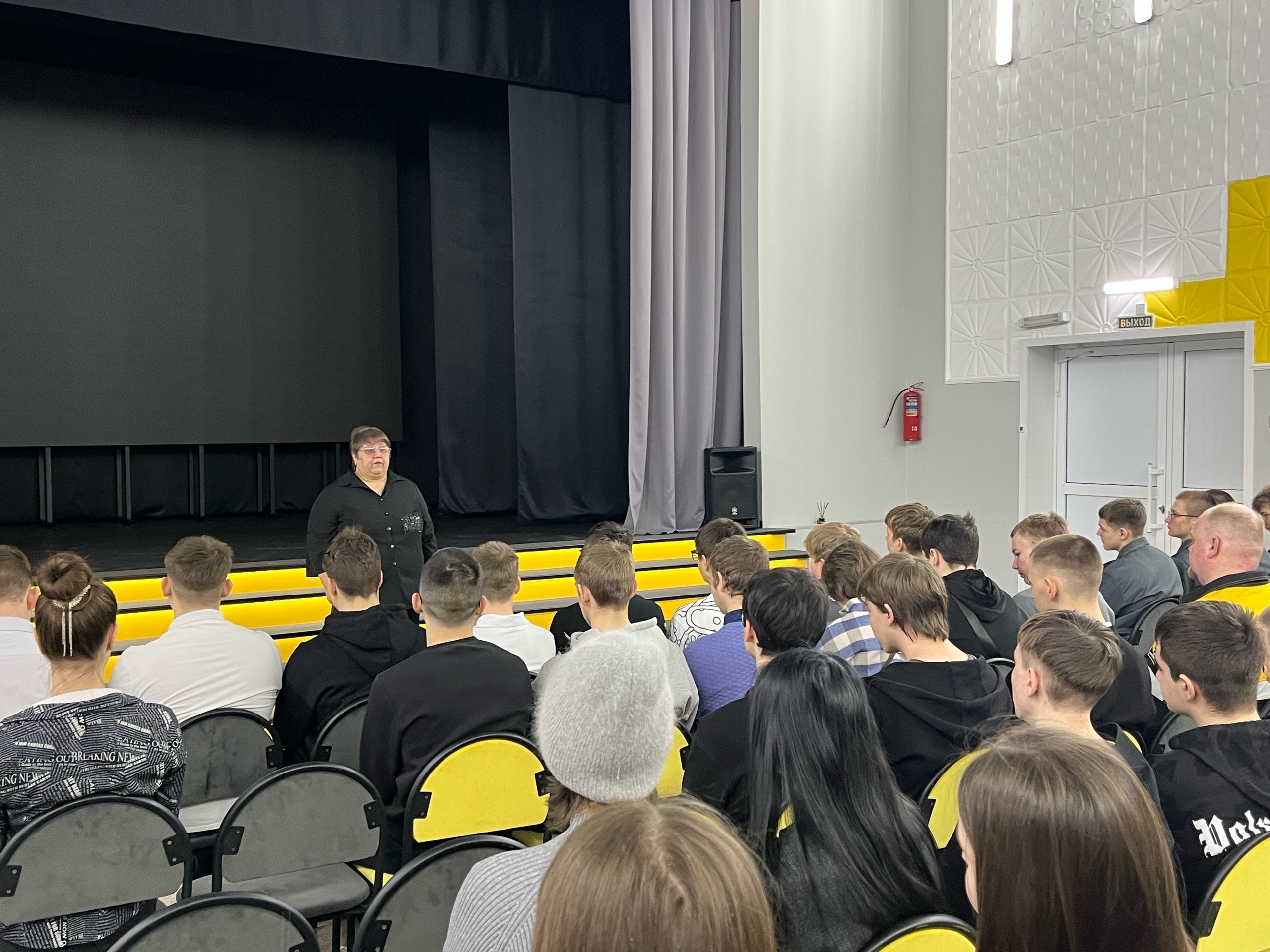 группу молодых людей, сидящих в аудитории. В центре расположена сцена с черной занавеской. Спикер (возможно, преподаватель или тренер) стоит спиной к аудитории и говорит с ней. Присутствующие одеты в повседневную одежду, в основном в темных тонах. На заднем плане можно заметить желтые скамейки, а освещение делит пространство на светлые и тенистые участки.