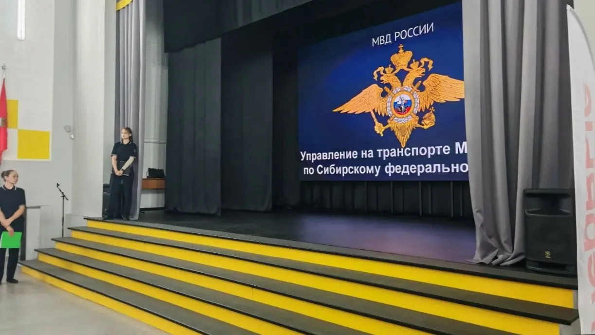 Общий план сцены актового зала. На заднем плане сцены расположен большой светящийся экран, на котором изображен герб МВД России (золотой двуглавый орел на синем фоне). Текст на экране гласит: сверху — «МВД РОССИИ», снизу — «Управление на транспорте М... по Сибирскому федерально...». Слева на сцене стоит женщина в темно-синей форме, руки сложены перед собой. Слева внизу у сцены стоит еще одна женщина в такой же форме, держащая зеленую папку. Сцена имеет широкие желтые ступени. Слева на стене видны флаги (красный и желто-белый), справа на сцене стоит черная аудиоколонка. Задний план сцены закрыт темными шторами.