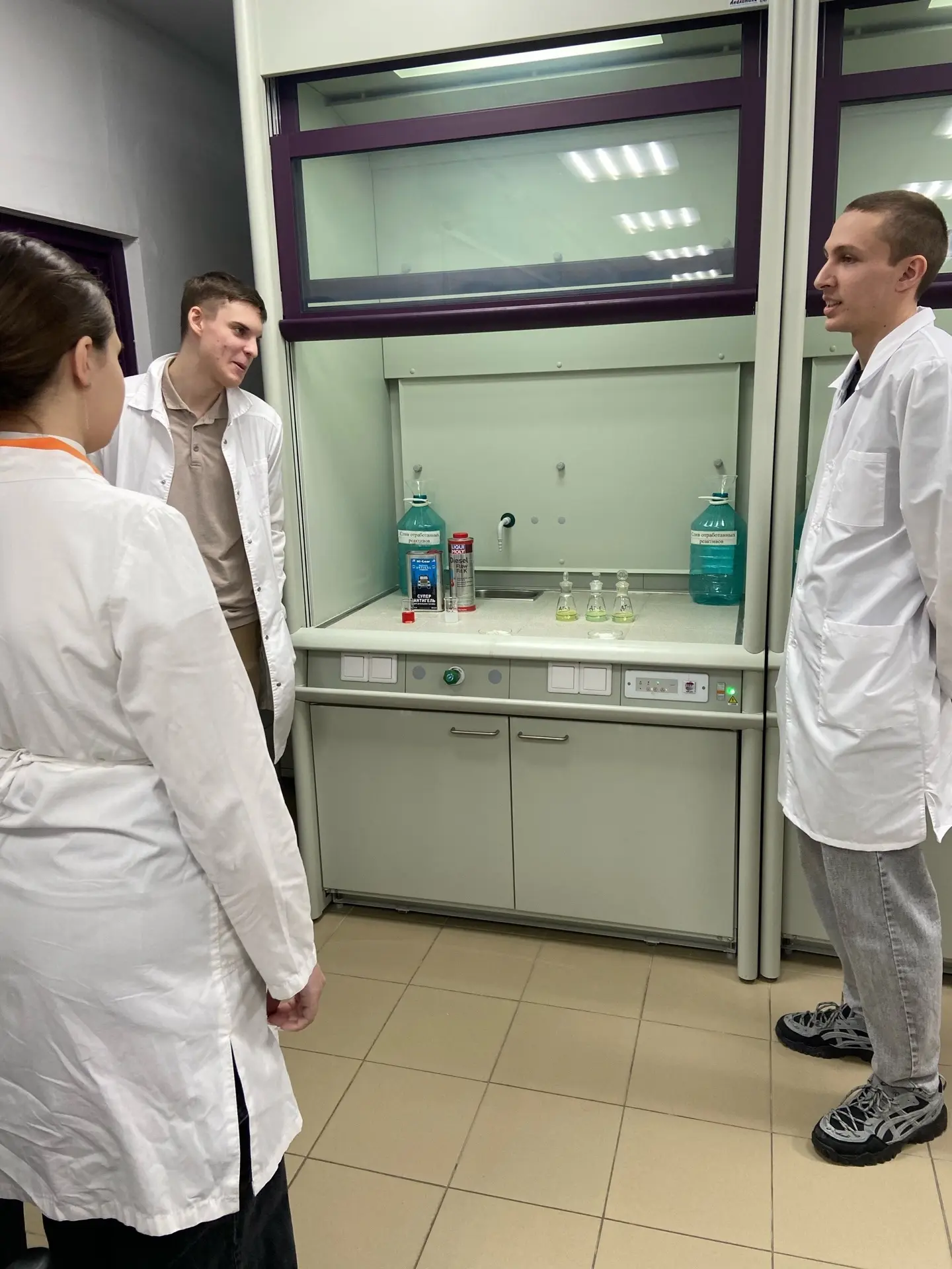 На фотографии двое молодых людей, стоящих у лабораторного оборудования, и третья девушка, стоящая немного позади них. Все они в белых халатах. Оборудование включает вытяжной шкаф с флаконами на столе. Мужчины общаются друг с другом, а девушка слушает. Пол желтого цвета, а стены серые, с ярким освещением.