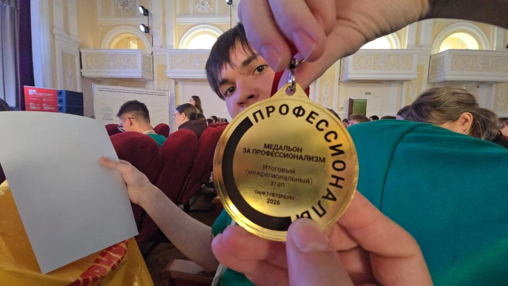 На фото изображён юноша, сидящий в аудитории, с медалью в руках. Медаль золотого цвета с надписью на чёрном фоне, которая гласит: "МЕДАЛЬОН ЗА ПРОФЕССИОНАЛИЗМ". Юноша с короткими тёмными волосами и выразительными чертами лица. Он одет в зелёную футболку и смотрит на камеру, улыбаясь. В зале видно много людей сзади, на фоне – светлые стены.