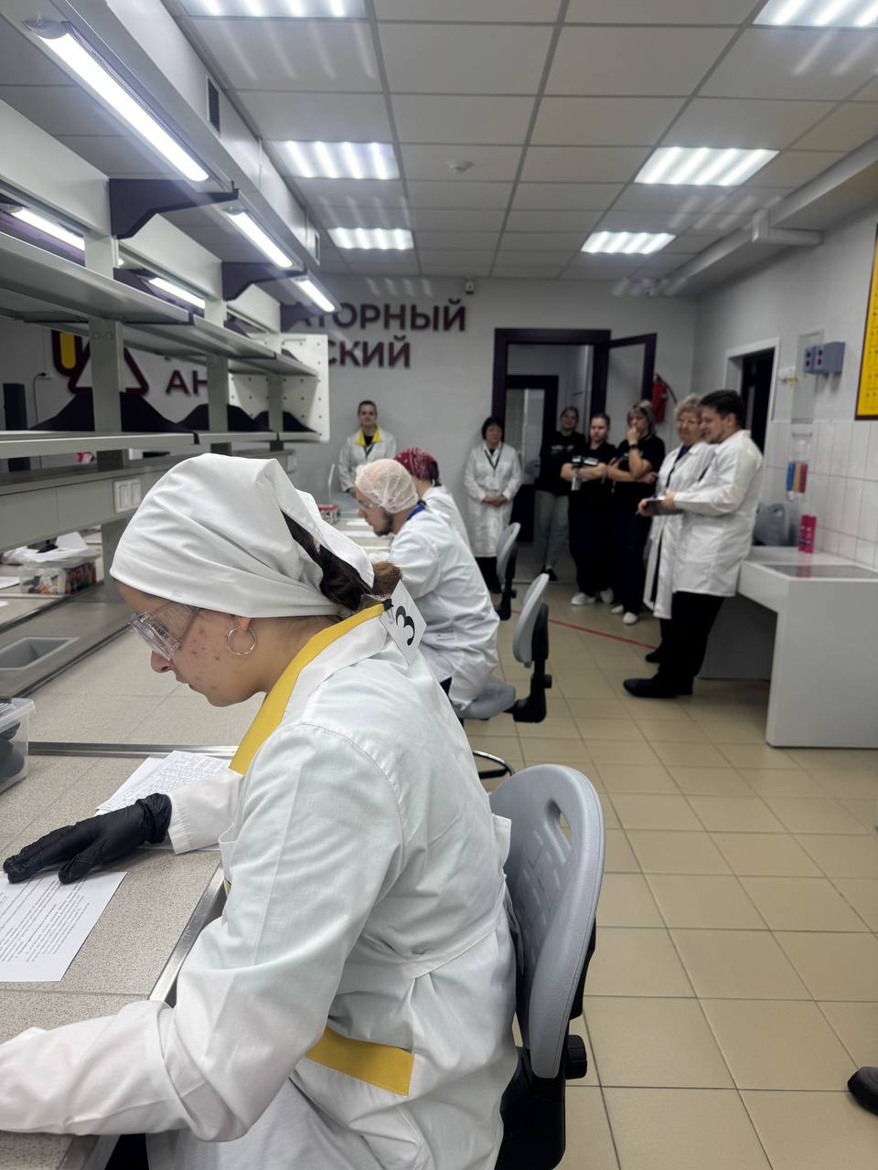 представлена лаборатория. Здесь видно, как две женщины в лабораторных халатах и защитных очках занимаются написанием заметок. В фоновом плане стоит группа людей, наблюдающая за их работой. Все участники учебного процесса сосредоточены, атмосфера настроена на обучение и обмен знанием. Стены окрашены в светлые тона, что придает помещению просторный вид.