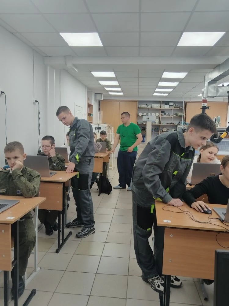 Класс с рядами парт: подростки за ноутбуками и взрослые наставники помогают им работать за компьютерами.
