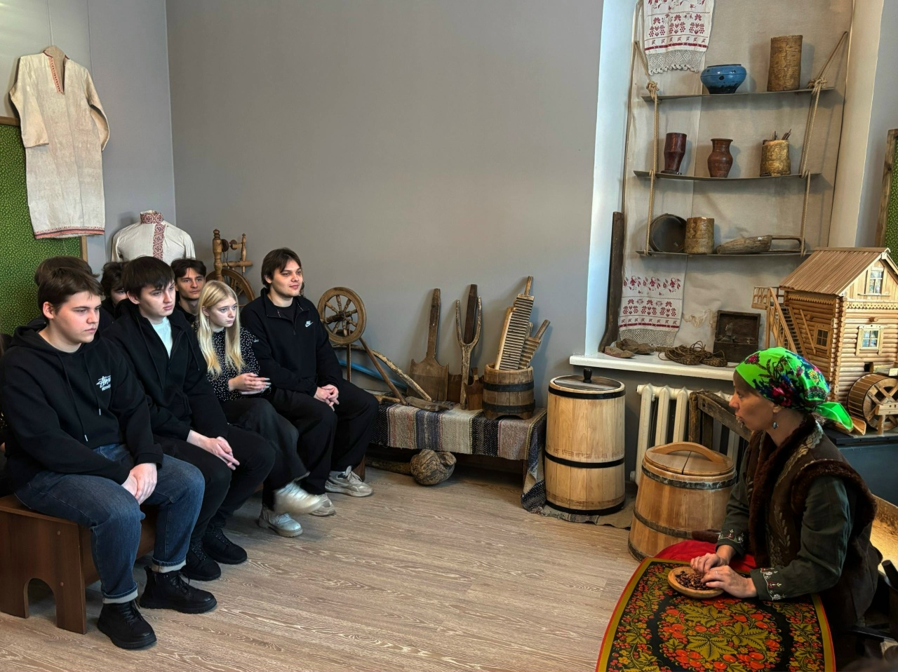 изображена другая группа молодежи, состоящая из шести человек, которые сидят на скамейке и внимательно слушают женщину, сидящую против них. Женщина одета в традиционный русский народный костюм с ярким платком. За ней находятся предметы старинного быта, такие как домашняя утварь и инструменты. Стены оформлены в нейтральных тонах, а пол выполнен из светлого дерева. Атмосфера учебная и дружелюбная, создается ощущение погружения в местную культуру.