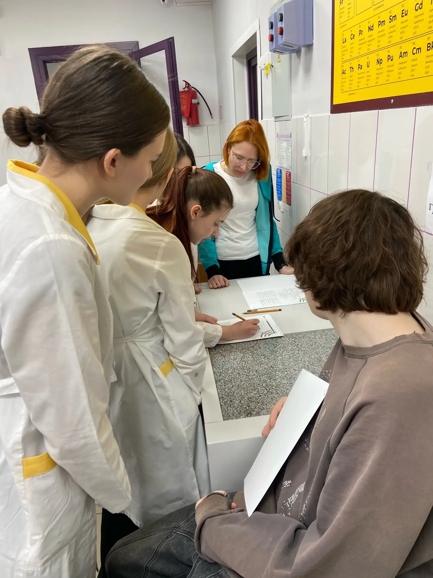 На изображении группа из пяти девушек, собравшихся за столом в лаборатории. Они в белых халатах и внимательно о чем-то обсуждают. Одна из них, стоящая слева, заполняет бумагу, а остальные смотрят на неё. На стене видны плитки с химическими элементами. Освещение яркое, а поверхности в помещении чистые.