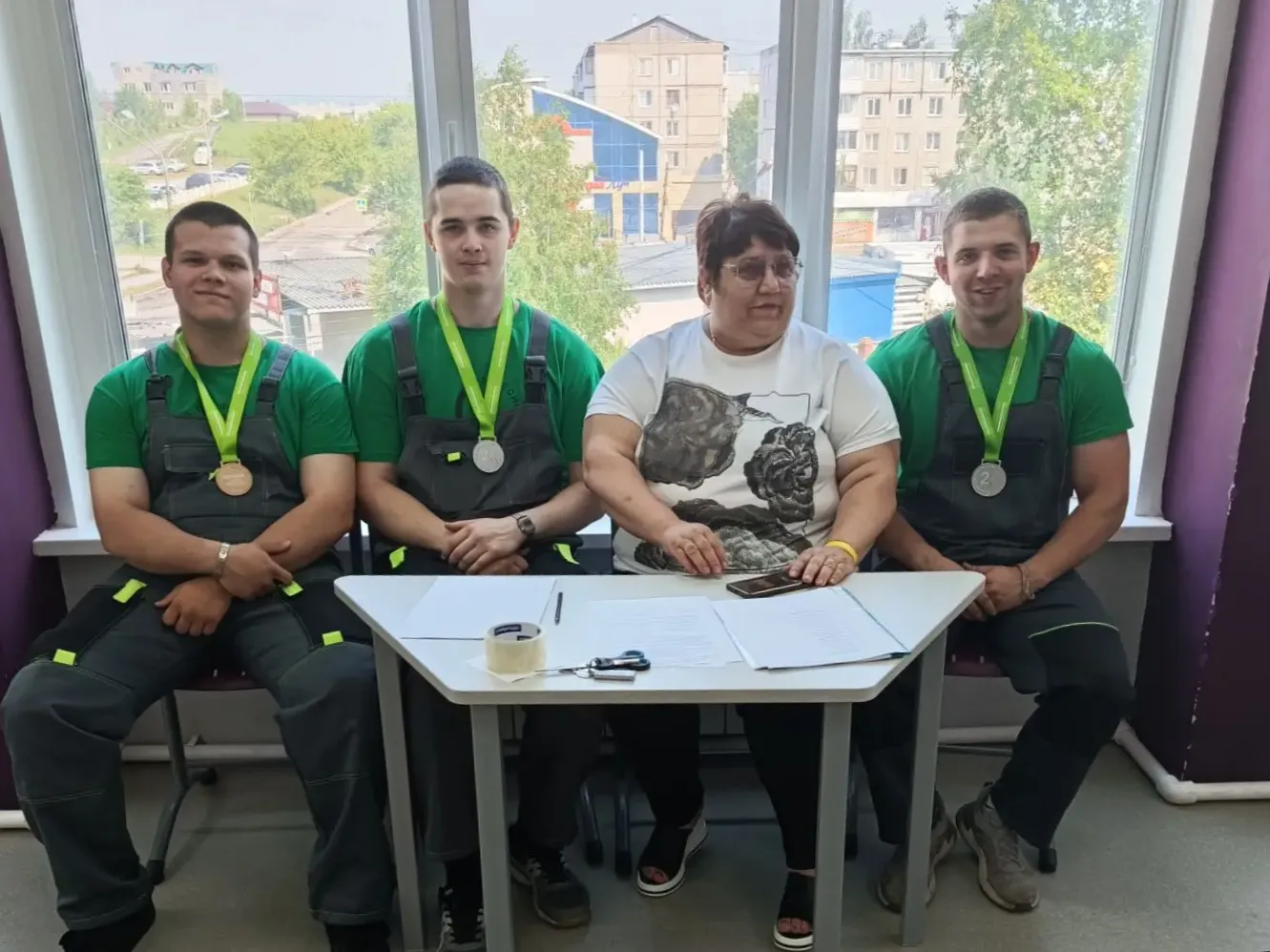 Группа из четырёх человек сидит у окна: три молодых мужчины в рабочей форме и с медалями на лентах, женщина в белой футболке сидит в центре за столом с бумагами и скотчем.