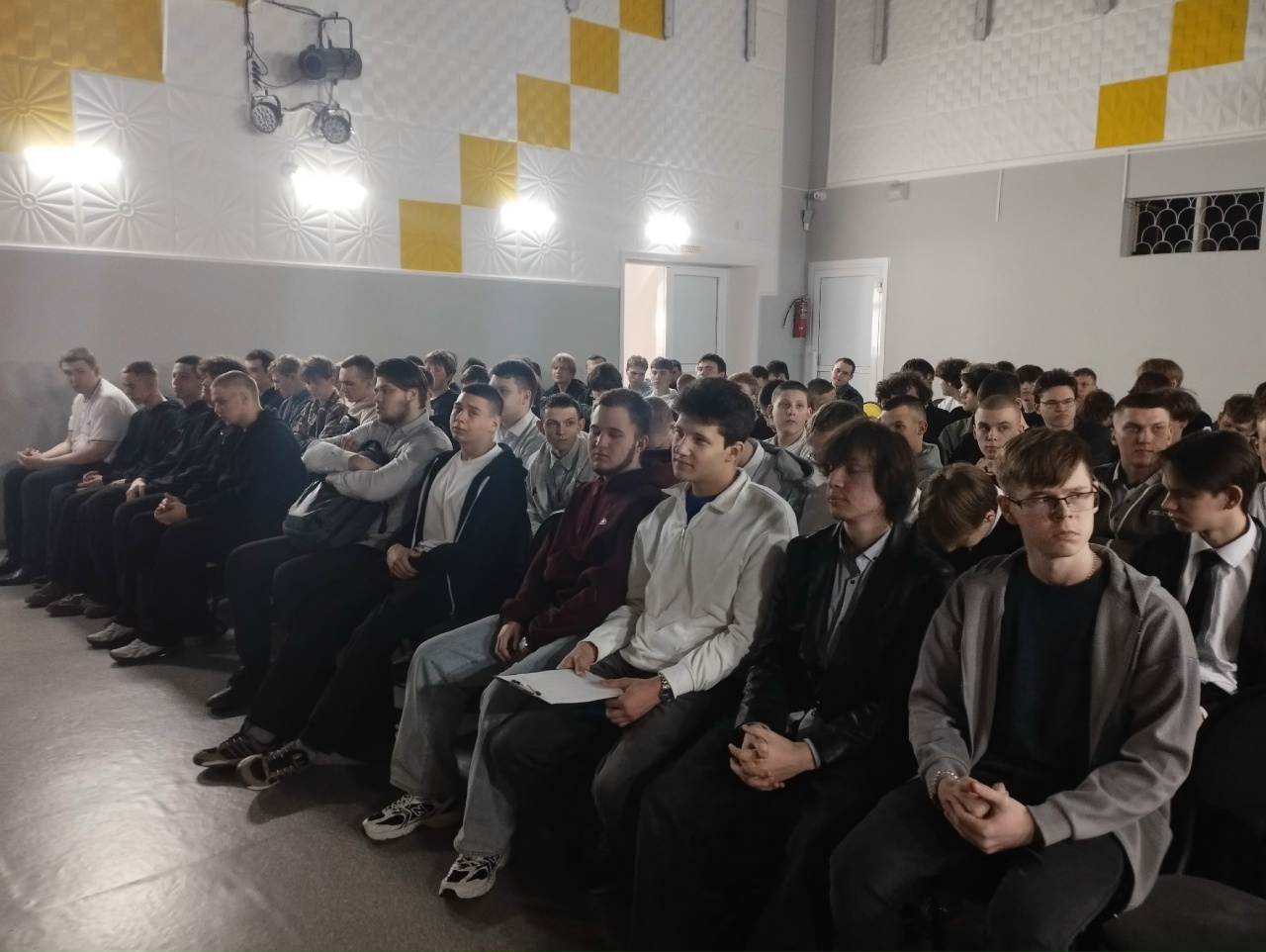 запечатлена большая группа молодых людей, сидящих в аудитории. Они внимательно смотрят вперед, большинство из них в белых или темных рубашках. Аудитория оформлена с использованием светлых и темных тонов, а освещение создает атмосферу сосредоточенности. Видно, что они собрались для совместного просмотра или обсуждения важного мероприятия.