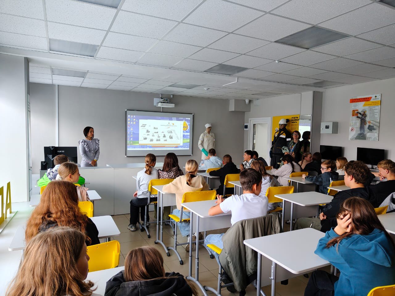 Это изображение внутреннего класса с учениками, сидящими за столами. На переднем плане две женщины, одна из которых стоит, а другая улыбается и демонстрирует информацию на экране проектора. Ученики, одетые в различные повседневные одежды, внимательно слушают и смотрят на экран. Комната оформлена в светлых тонах, с освещением, создающим уютную атмосферу.
