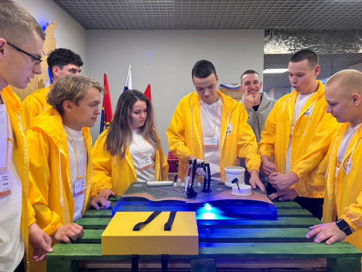 Группа подростков в ярко-желтых куртках стоит вокруг стола, на котором расположена модель какого-то устройства или станции. Участники сосредоточенно смотрят на модель, обсуждая детали. На фоне видно многоцветные флаги и элементы экспозиции.